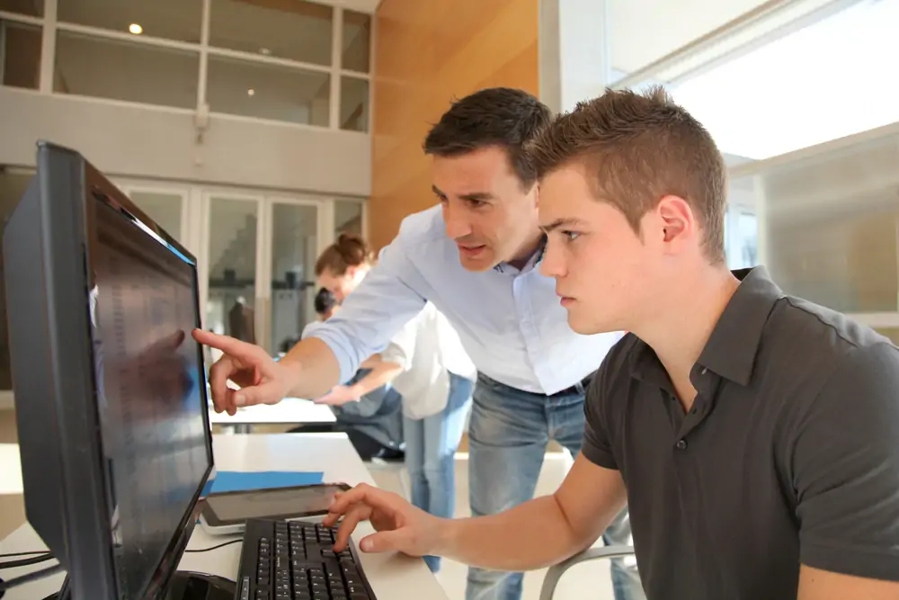 Ein Fachinformatiker in der Ausbildung an einem PC mit dem Lehrer neben sich