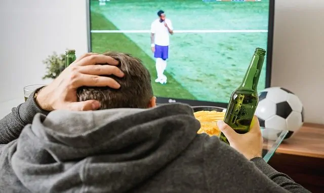 Ein junger Mann sitzt mit einer Bierflasche in der Hand vor dem Fernseher und schaut Fußball
