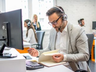 Ein junger Mann im Anzug sitzt mit Headset vor dem PC am Arbeitsplatz