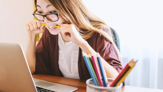 Eine junge Frau mit Brille beißt auf einen Stift und sitzt vor einem Notebook