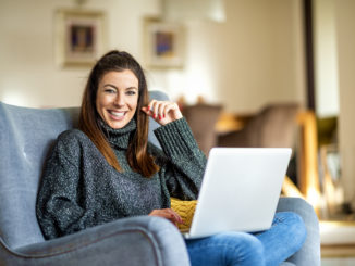 Eine junge Dame mit braunen Haaren sitzt mit einem weißen Notebook auf einem blauen Sessel.